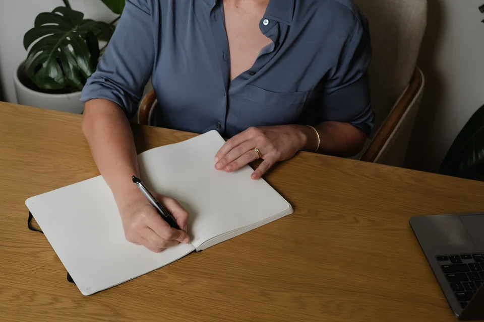 Close-up of Annie Wright writing in an open notebook at a wooden desk, with her laptop beside her.