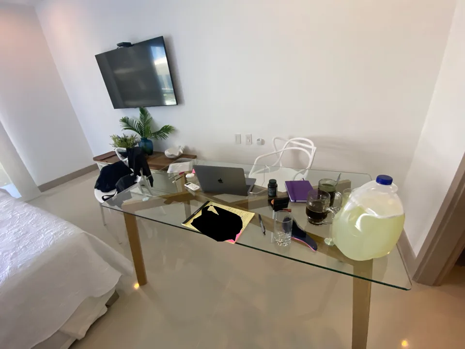 A laptop and legal pad on a glass table beside a bed, with a pitcher of lemonade. The original image includes a redacted area on the pad.