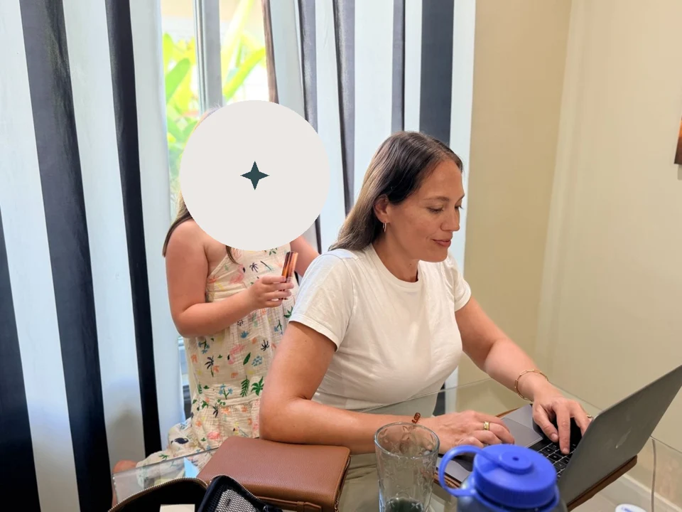Annie working at her laptop while her daughter brushes her hair. Her daughter's face is covered by an opaque privacy mark.