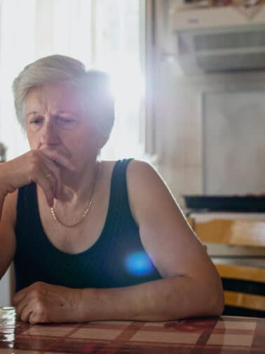 Senior woman looking unhappy sitting alone in the kitchen wondering, Why am I so negative?
