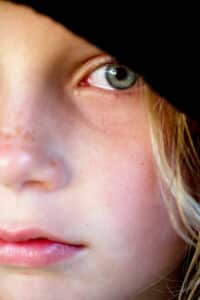 Image of off centered macro portrait of young blonde girl crying. She looks very sad. She has a black hood on her head, freckles on her nose and rosy lips representing childhood trauma despite a materially privileged background