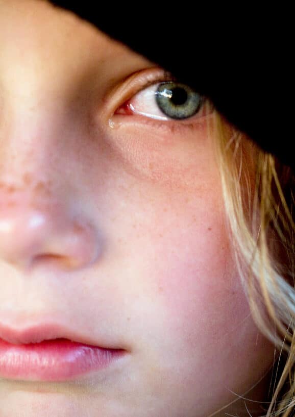 Image of off centered macro portrait of young blonde girl crying. She looks very sad. She has a black hood on her head, freckles on her nose and rosy lips representing childhood trauma despite a materially privileged background