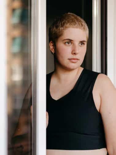 Young woman with short hairstyle in black top standing near an open window and looking out wondering What is trauma and what impacts might have had on her.