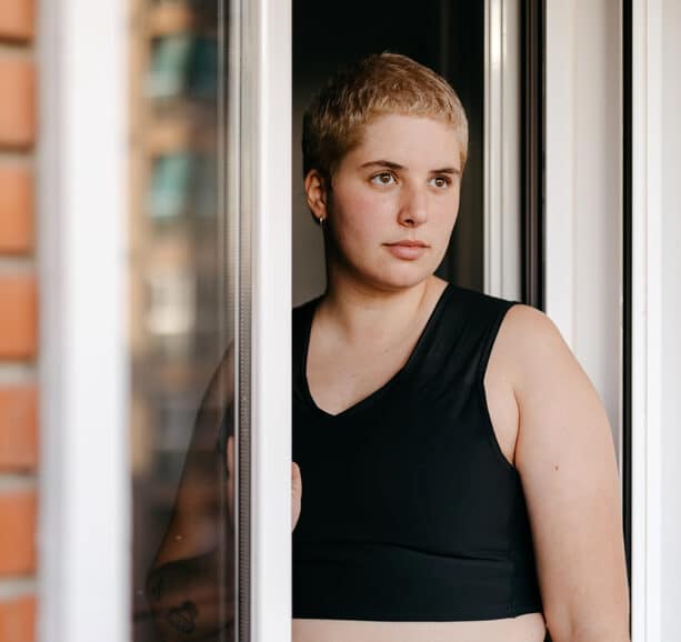 Young woman with short hairstyle in black top standing near an open window and looking out wondering What is trauma and what impacts might have had on her.