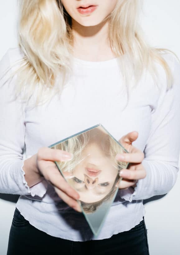 Photo of a casual model looking at camera though sharp piece of broken mirror holding in hands representing the relational trauma definition