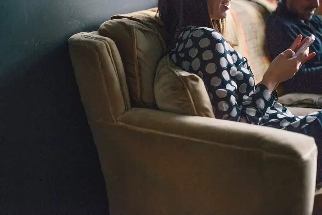 A woman sits on a couch looking at her phone beside another person, capturing the quiet distance and disconnection that can linger from childhood trauma.
