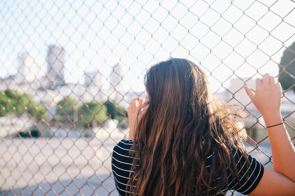 A woman standing behind a chain-link fence, symbolizing the trapped and conflicted feelings often experienced in trauma bonding.