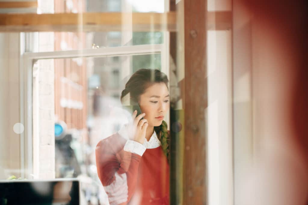 A person stands by a window talking on the phone, their thoughtful expression suggesting emotional tension and the nervous system’s response to stress or trauma.